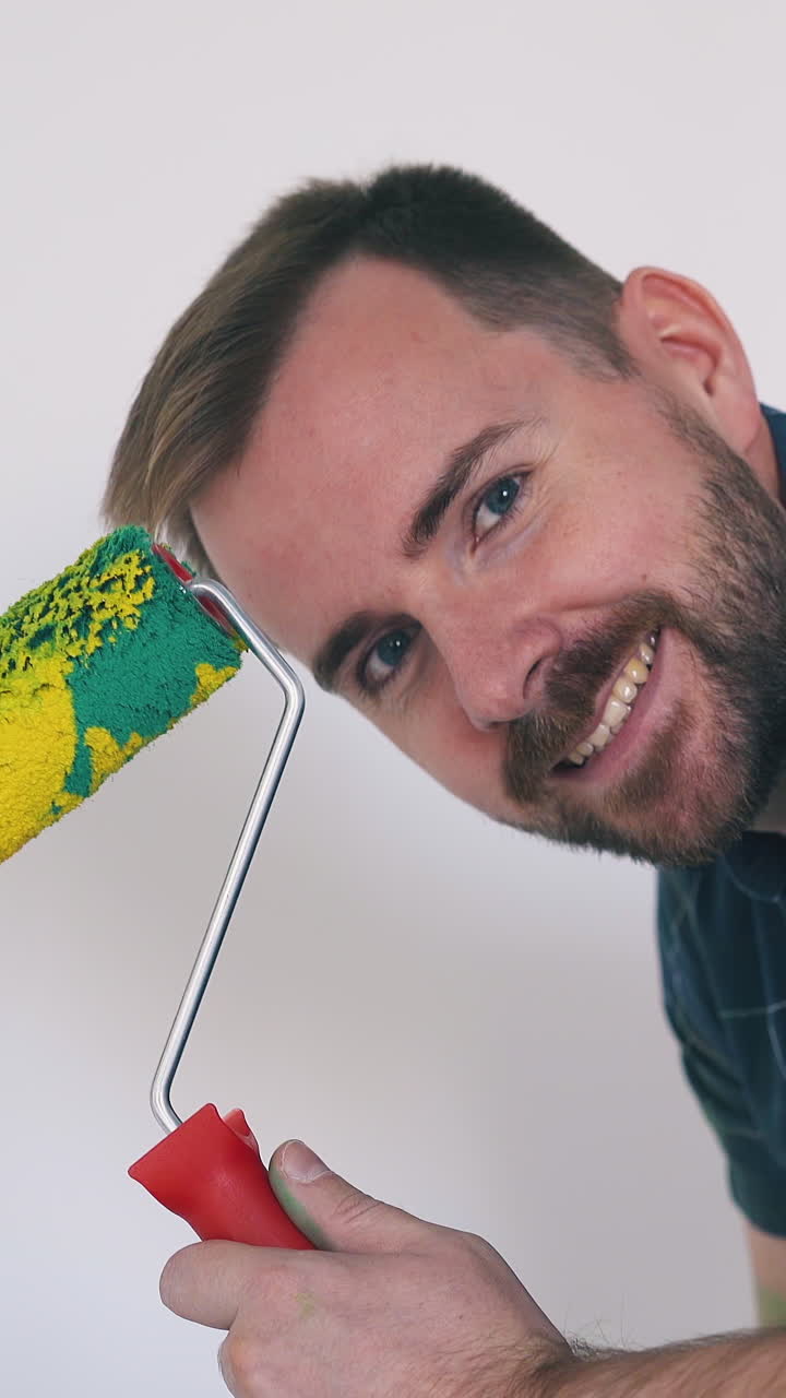 joyful blue eyed builder in dark shirt shows roller with bright yellow paint against white wall in repairing room closeup