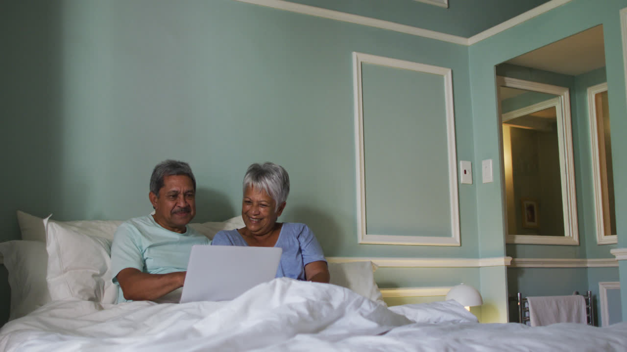 Happy senior mixed race couple sitting in bed using laptop