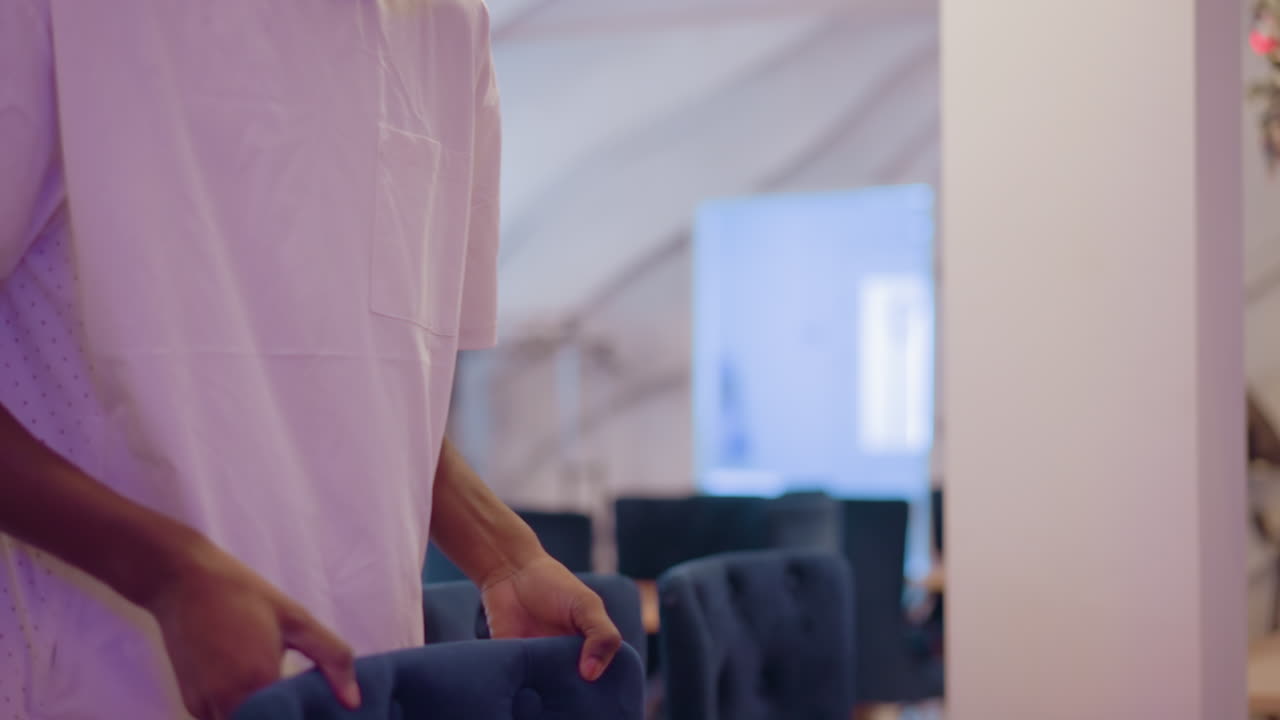 Person in white shirt carefully adjusting blue tufted chairs in bright, modern room, preparing space for an event or gathering, with focus on organizing furniture arrangement