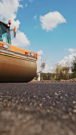 Orange roller machine pressing hot asphalt on the road in summer. Heavy compactor machine making new asphalt on the background of blue sky and green trees. Road resurfacing. Vertical video