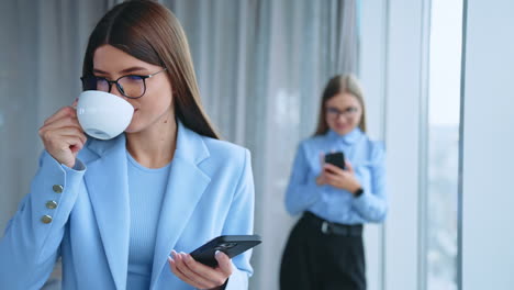 beautiful dark-haired young female wearing glasses drinking coffee looking at her phone. female colleague messaging someone on smartphone at backdrop in blur.