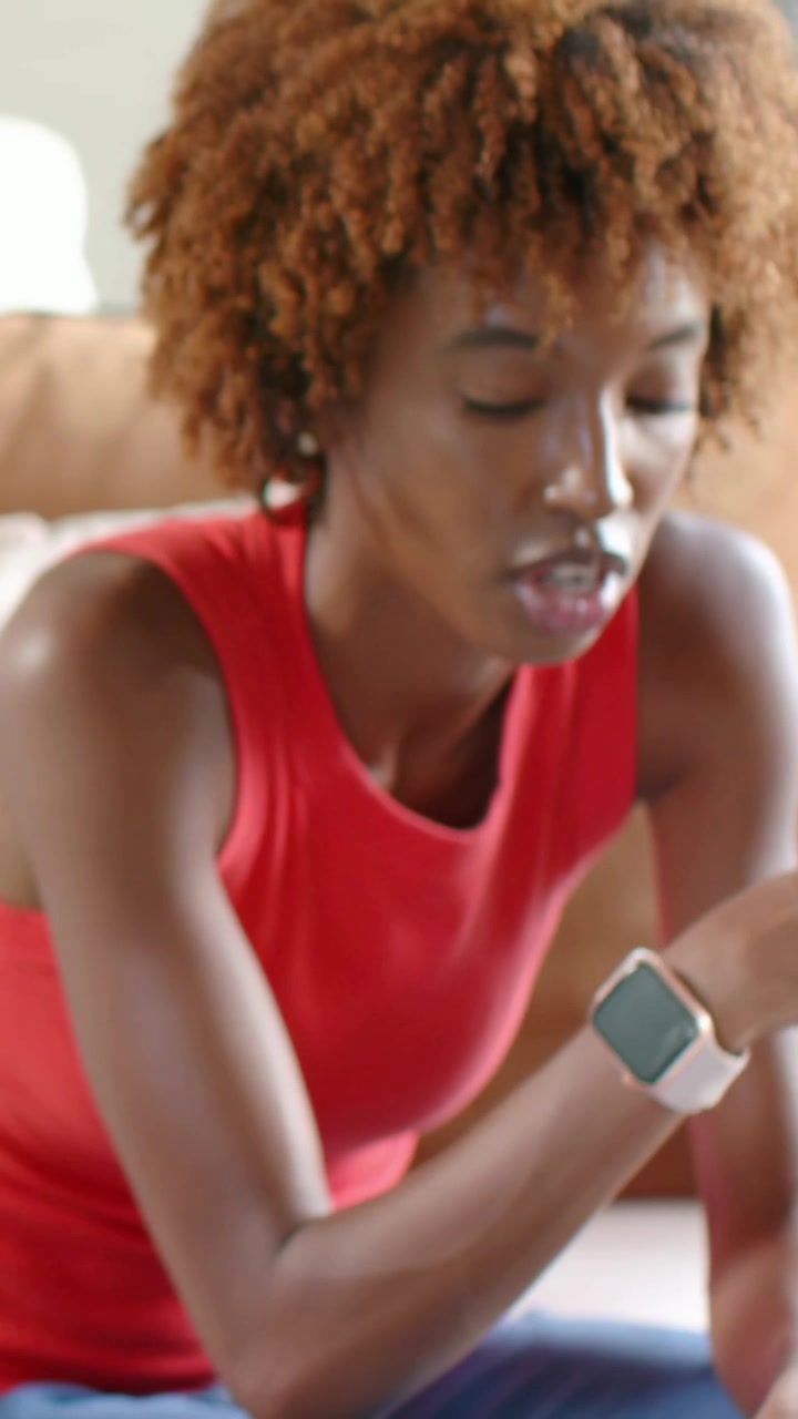 Vertical video: Camera focusing woman in red top, jeans leaning on sofa, checking laptop and watch