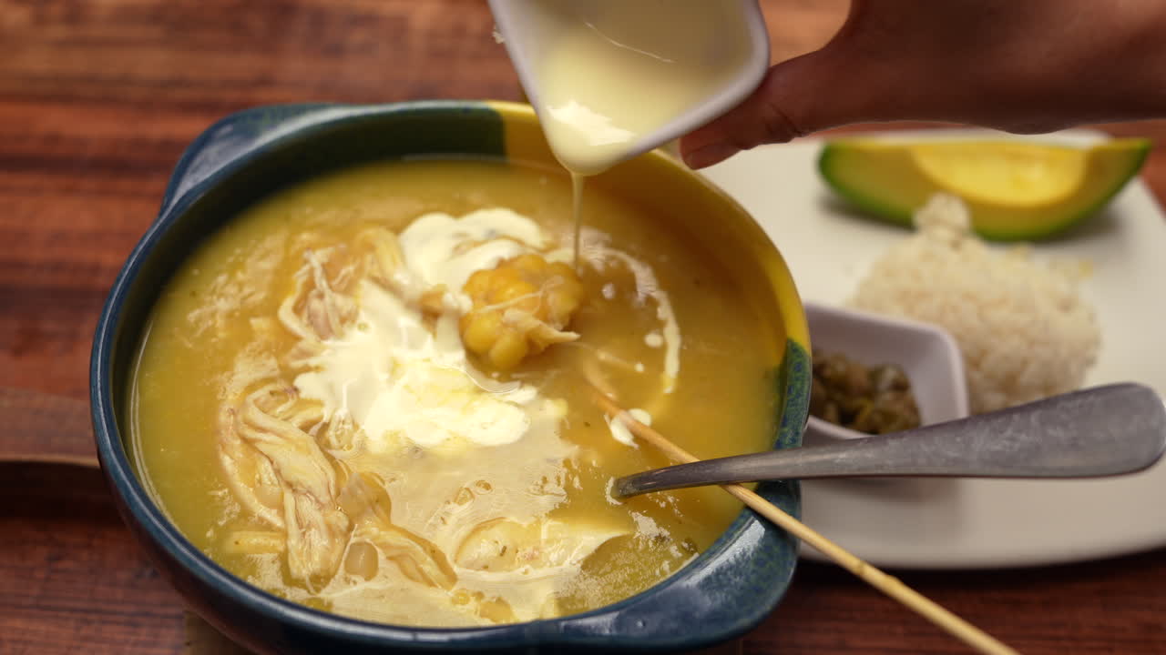 Detailed close-up shot of traditional Colombian ajiaco soup with creamy topping. Warm lighting enhances rich textures. Hand pouring cream adds a homely touch to this culinary scene