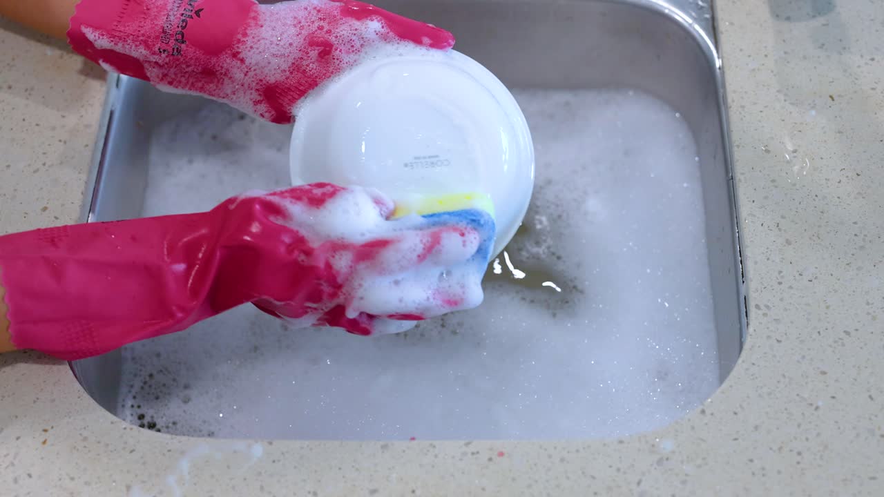 Hands in pink gloves wash a dish in a soapy sink. Bright lighting highlights the clean, organized kitchen environment