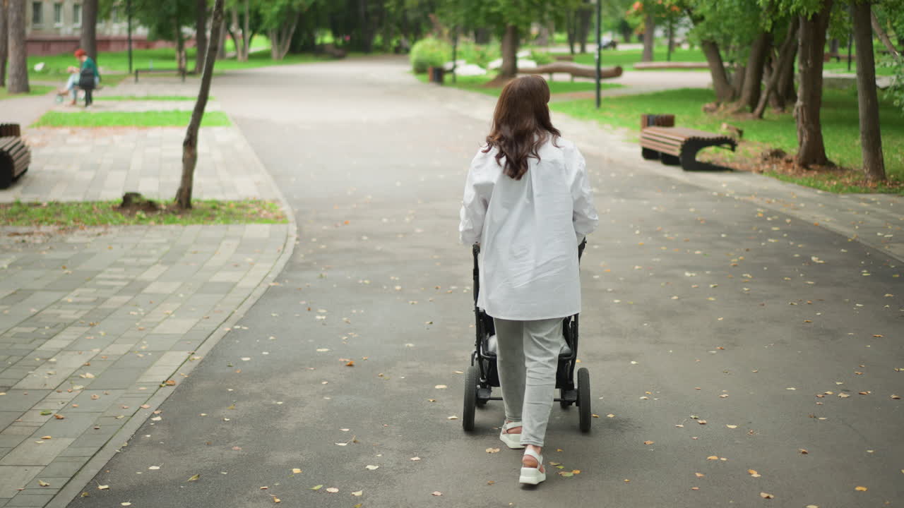 Kvinne med barnevogn. Stille morgentur med barnevogn. En kvinne spaserer fredelig med barnevogn i parken. Kvinne går rolig med barnevognen sin langs en stille og bladrik parksti.