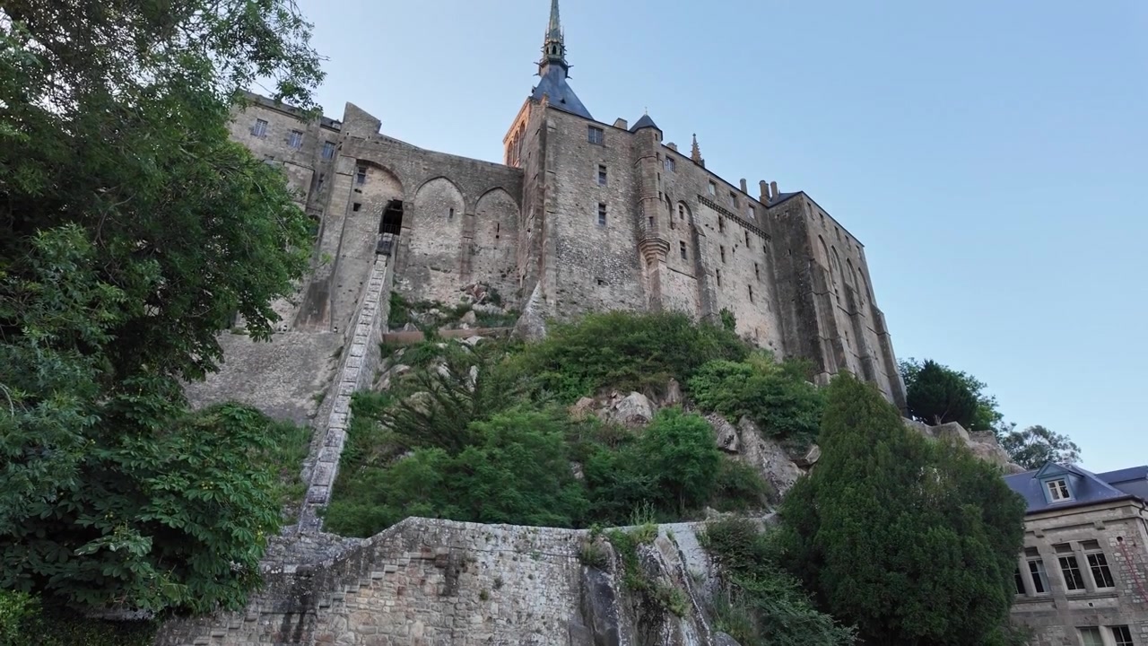 Vista laterale dell'Abbazia di Mont Saint-Michel, patrimonio UNESCO, che domina la sua base rocciosa, con mura medievali, vegetazione lussureggiante e un'impressionante architettura storica sotto un cielo sereno.