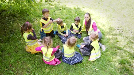 group little children sitting a circle with a tutor and playing games together a lawn in a park or forest. kindergarten a hike or trip. kids sit on the grass meadow have fun.