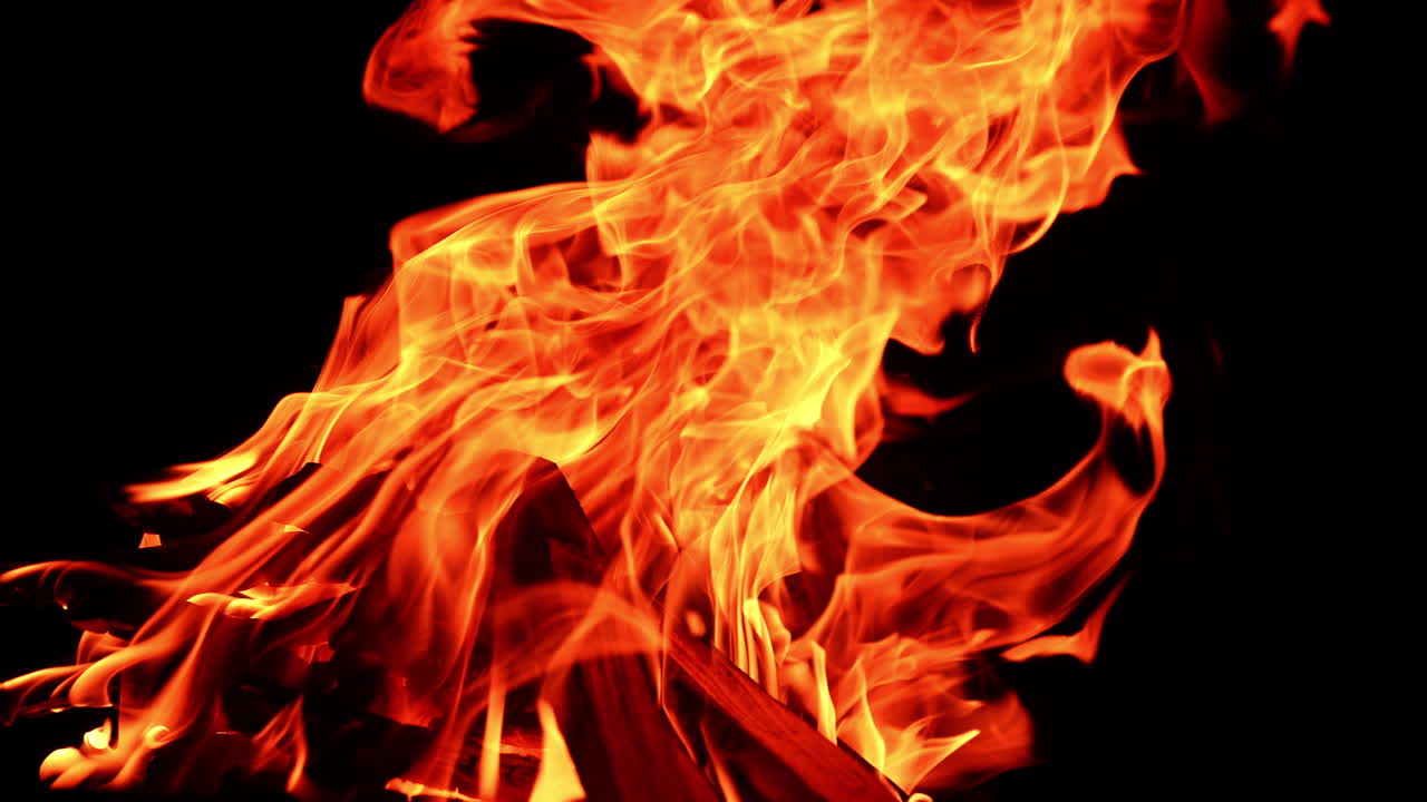 Flammen fra bålet strækker sig op og læner sig mod højre. En bunke træ brænder i lejrbålet. Nærbillede