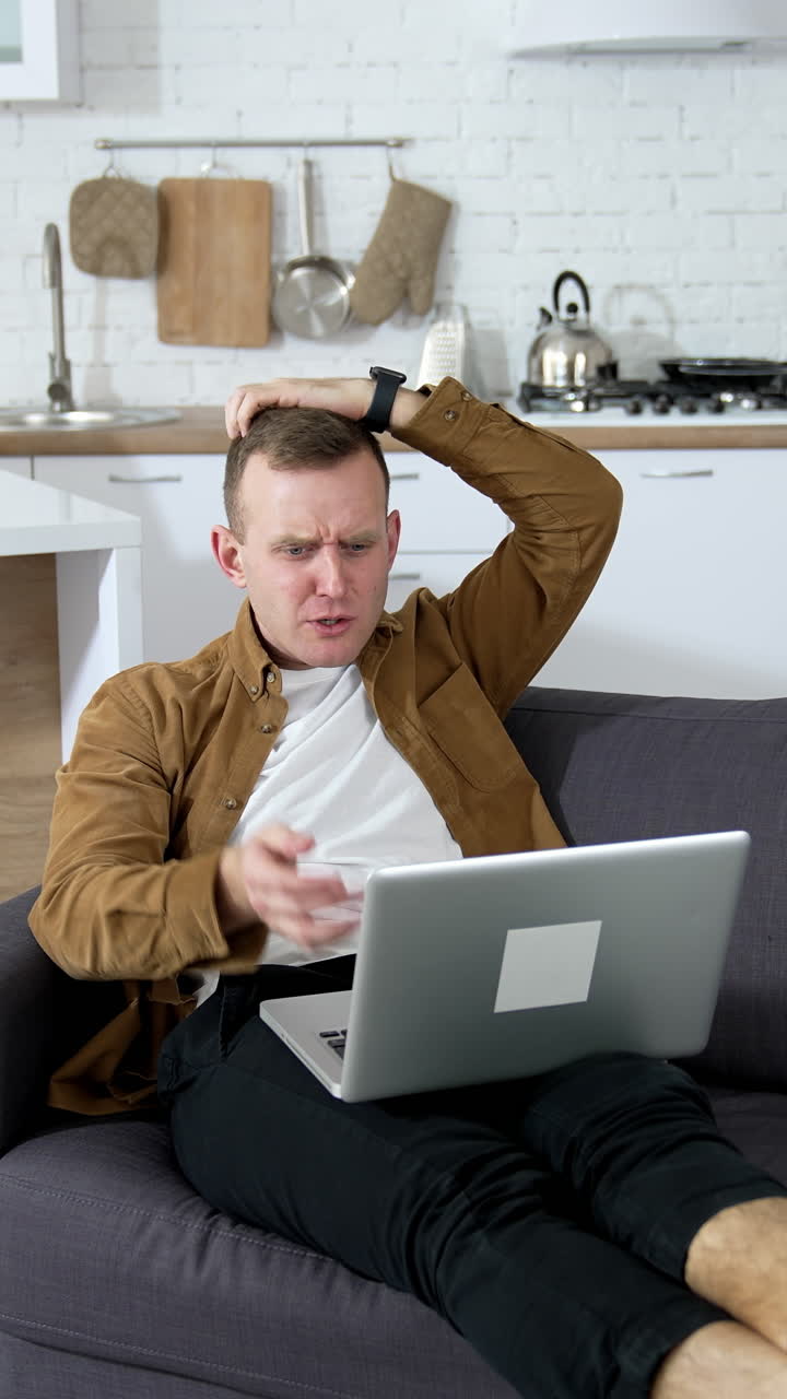 Young man playing computer games at home. Nervous guy laying on a couch with a laptop on kitchen background. Vertical video