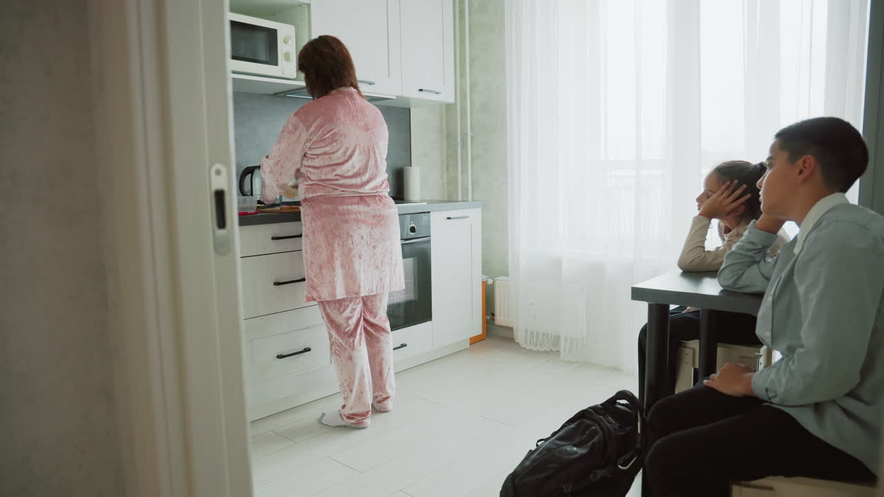 mother in pink pajamas cooking breakfast in bright modern kitchen two children sitting at table in school uniforms waiting impatiently with bags on floor morning routine scene familial