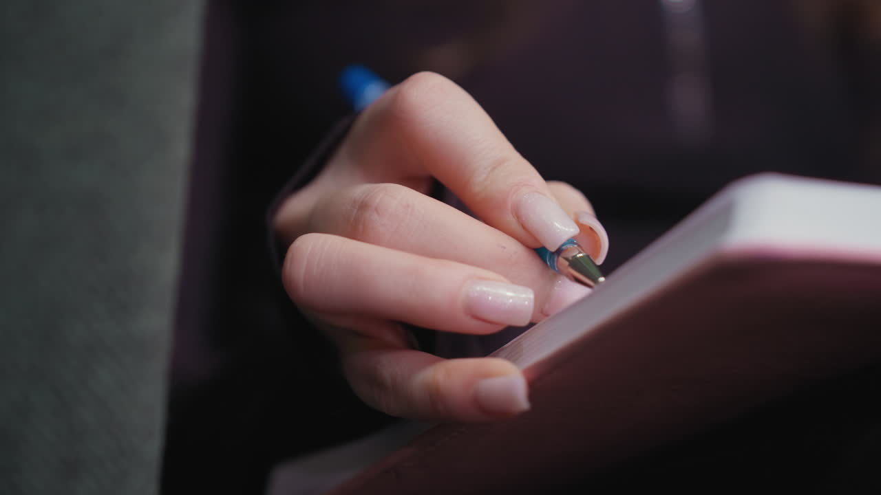 Close up of woman s hand with acrylic nails writing in pink notebook using blue pen, with detailed view of fingers and paper texture, showing focused motion in calm and creative indoor environment