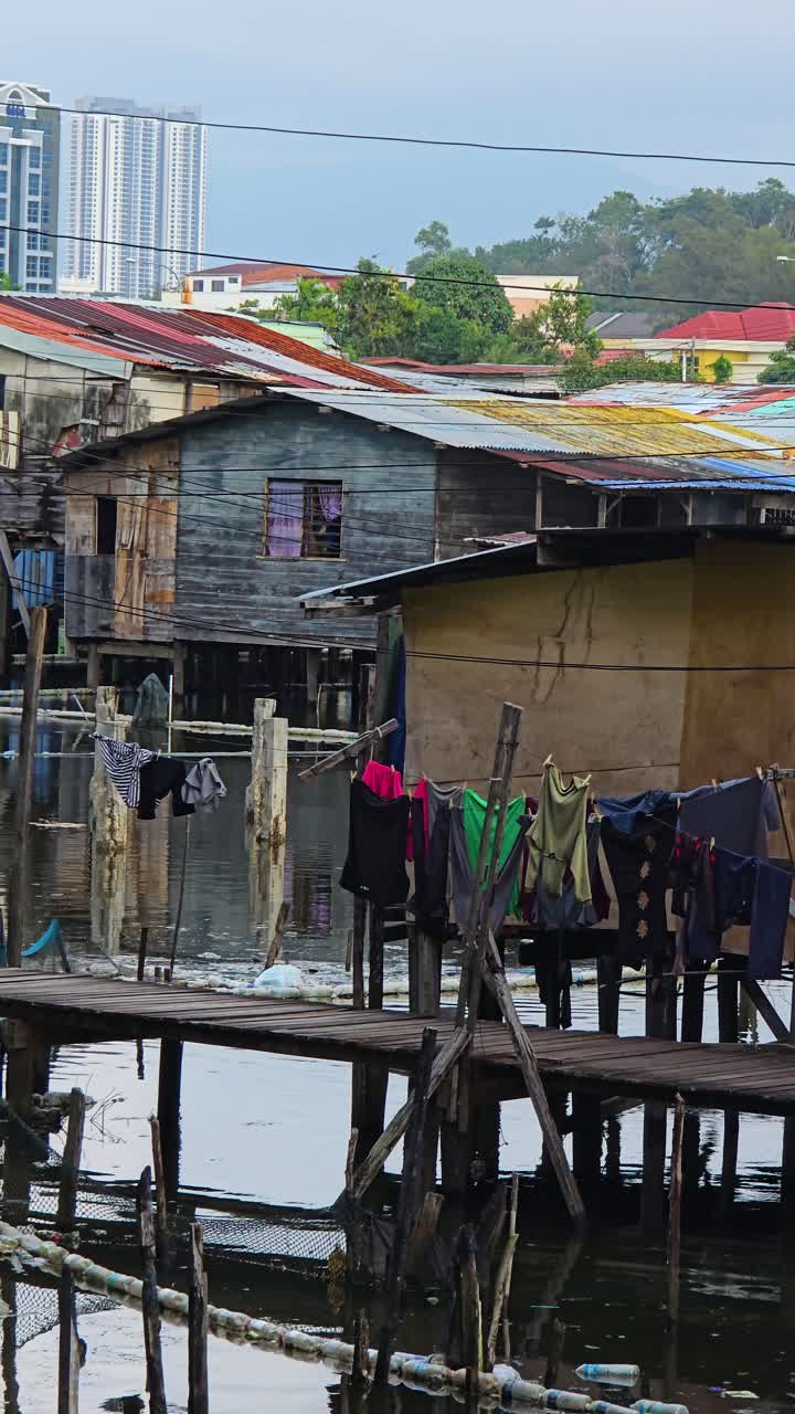View of a poor village built on stilts over water, with rustic wooden huts with metal roofs and laundry hanging to dry, against the backdrop of modern high-rise buildings in Kota Kinabalu, Sabah