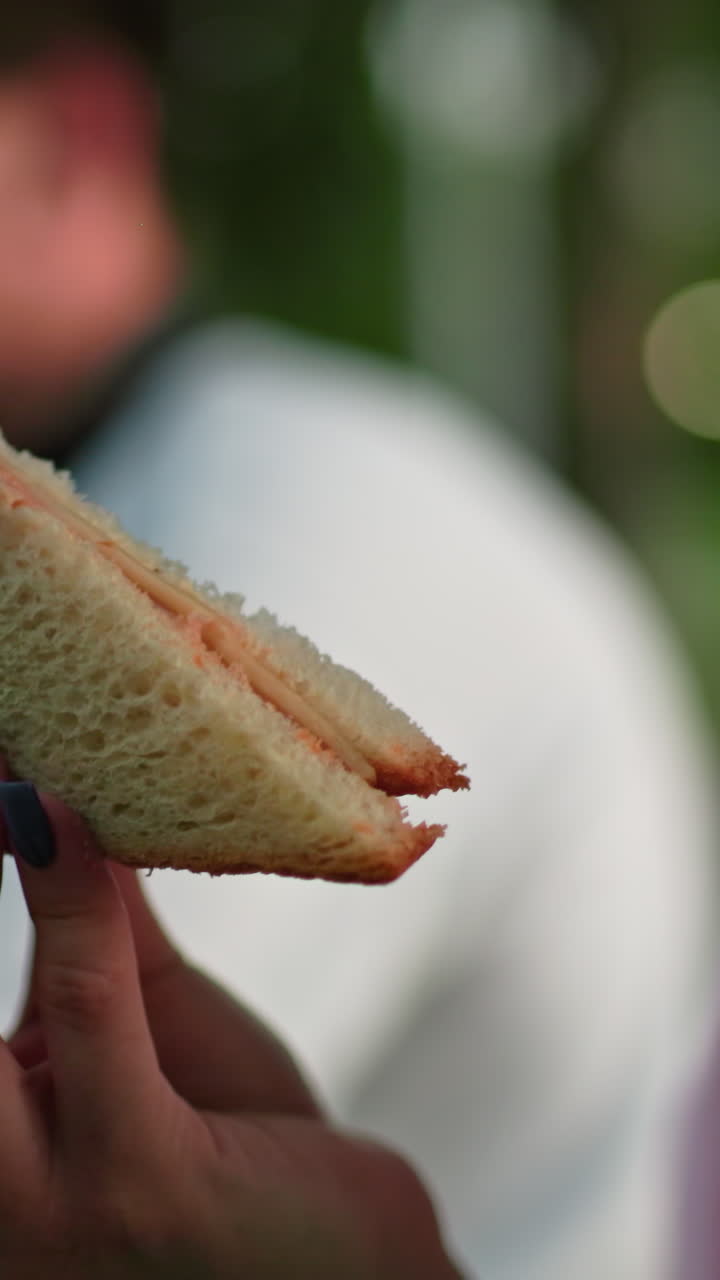 close-up de una mujer con uñas pulidas observando un sándwich a la parrilla en su mano, con un fondo borroso que muestra a alguien masticando sándwich en un entorno al aire libre con iluminación suave