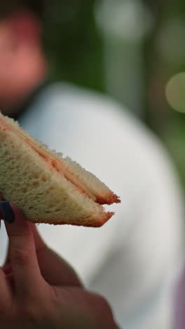 close-up de una mujer con uñas pulidas observando un sándwich a la parrilla en su mano, con un fondo borroso que muestra a alguien masticando sándwich en un entorno al aire libre con iluminación suave