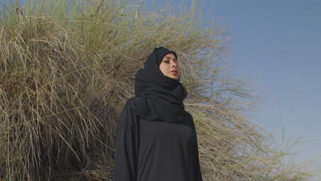 retrato de una hermosa mujer musulmana con vestido tradicional y hiyab de pie con los ojos cerrados cerca de un arbusto del desierto