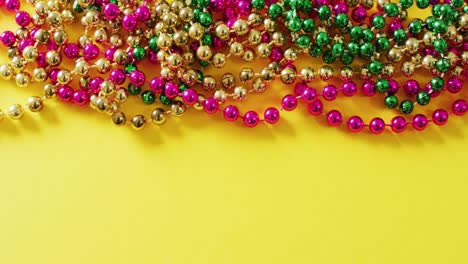 Video-of-pink,-gold-and-green-mardi-gras-carnival-beads-on-yellow-background-with-copy-space