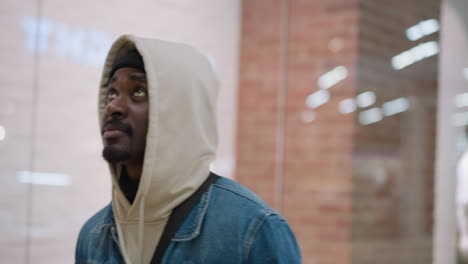 handsome young man wearing hoodie layered under denim jacket walking inside modern shopping mall, gazes thoughtfully at window display, urban casual style portrait with blurred mannequins and lights