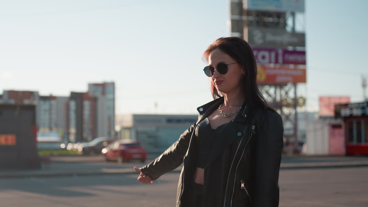Donna sicura di sé con giacca di pelle nera e occhiali da sole in piedi in una strada della città al tramonto che allunga il braccio con un gesto, esprimendo indipendenza, atteggiamento audace e stile di vita urbano alla moda