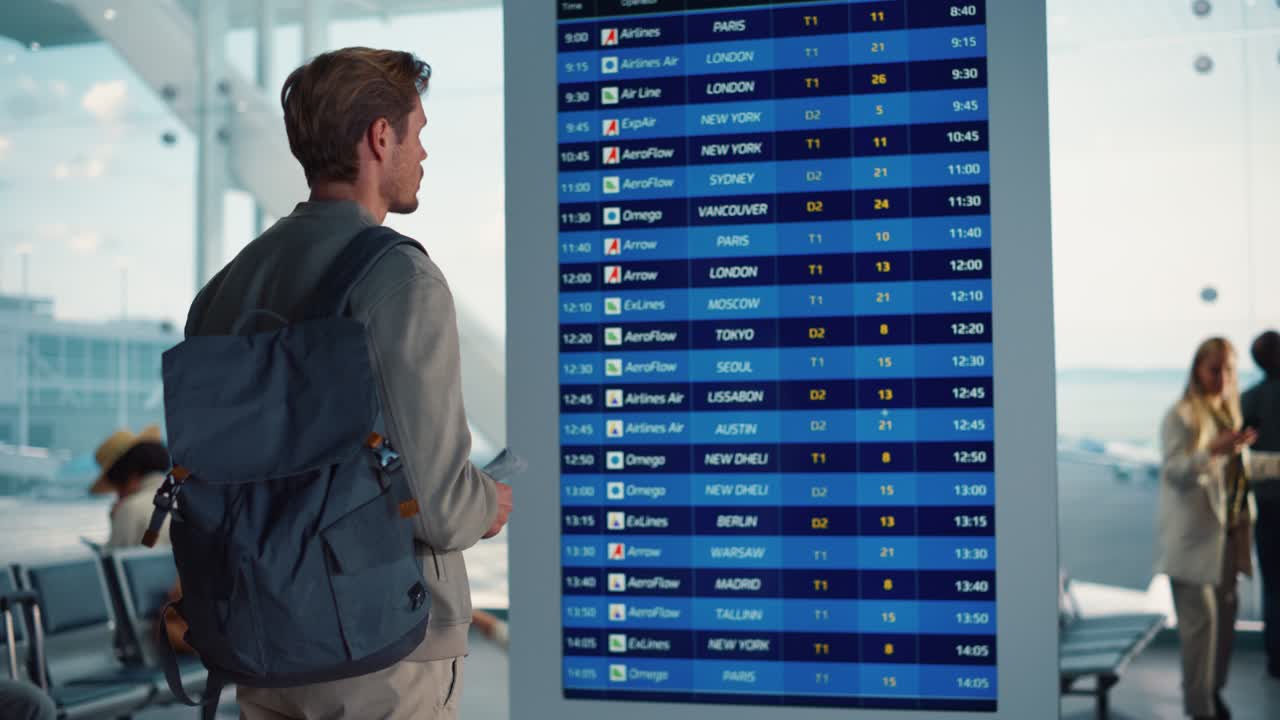 공항 터미널: 도착 및 출발 정보 디스플레이를보고있는 젊은이가 비행기를 찾고 있습니다. 배경: 다양한 사람들이 항공 허브의 탑승 라운지에서 비행기를 기다리고 있습니다.