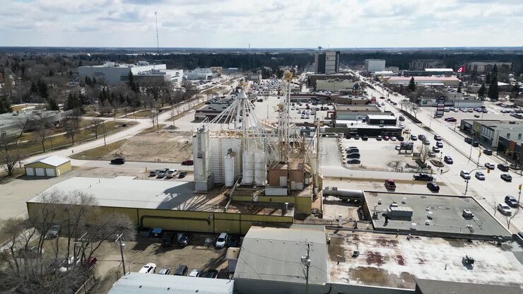 Lenta ripresa aerea cinematografica con drone che orbita attorno a un grande impianto di lavorazione industriale nel mezzo di una piccola città, con serbatoi di stoccaggio, fumo, strade vicine, persone ed edifici di piccole imprese visibili sotto
