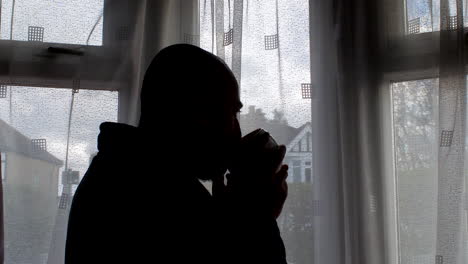 standing beside the window, silhouette of bald man savors his coffee while gazing at the daytime scene outside, exemplifying the essence of routine household moments