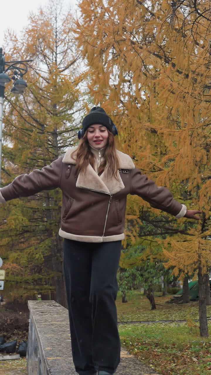 Student wearing headphone, black knit cap, brown shearling jacket, black trousers balancing gracefully with arms outstretched while walking joyfully on wide stone surface