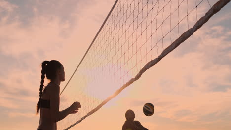 slow motion low angle close up lens flare: young female volleyball players pass and spike the ball over the net on a sunny summer evening. fit caucasian girls playing beach volleyball at sunset.