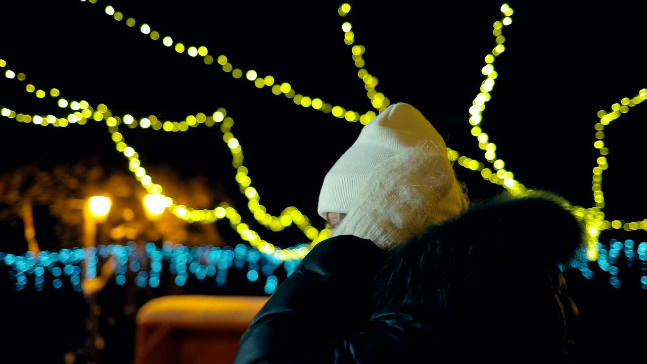 하 모자와 손바닥을 입은 소녀는 밤에 도시를 돌아다니며 미소 짓고 있습니다. 도시는 불빛으로 장식되어 있습니다. 카메라의 원형 움직임.