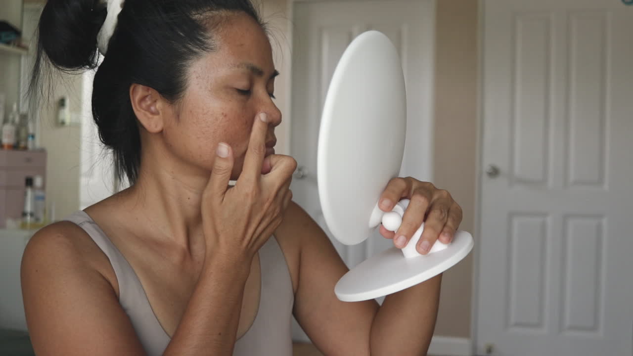 Giovane donna esamina l'acne usando uno specchietto a mano a casa, toccandosi il mento e osservando la condizione della pelle in un ambiente calmo e naturale.