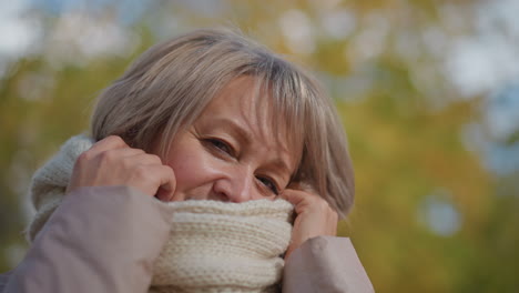 close up of woman clutching warm knitted scarf affectionately around neck with eyes closed, enjoying cozy outdoor moment in cool autumn weather with soft light and colorful blurred background