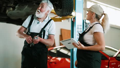 female mechanic and senior man worker using digital tablet standing under a car at a car service station