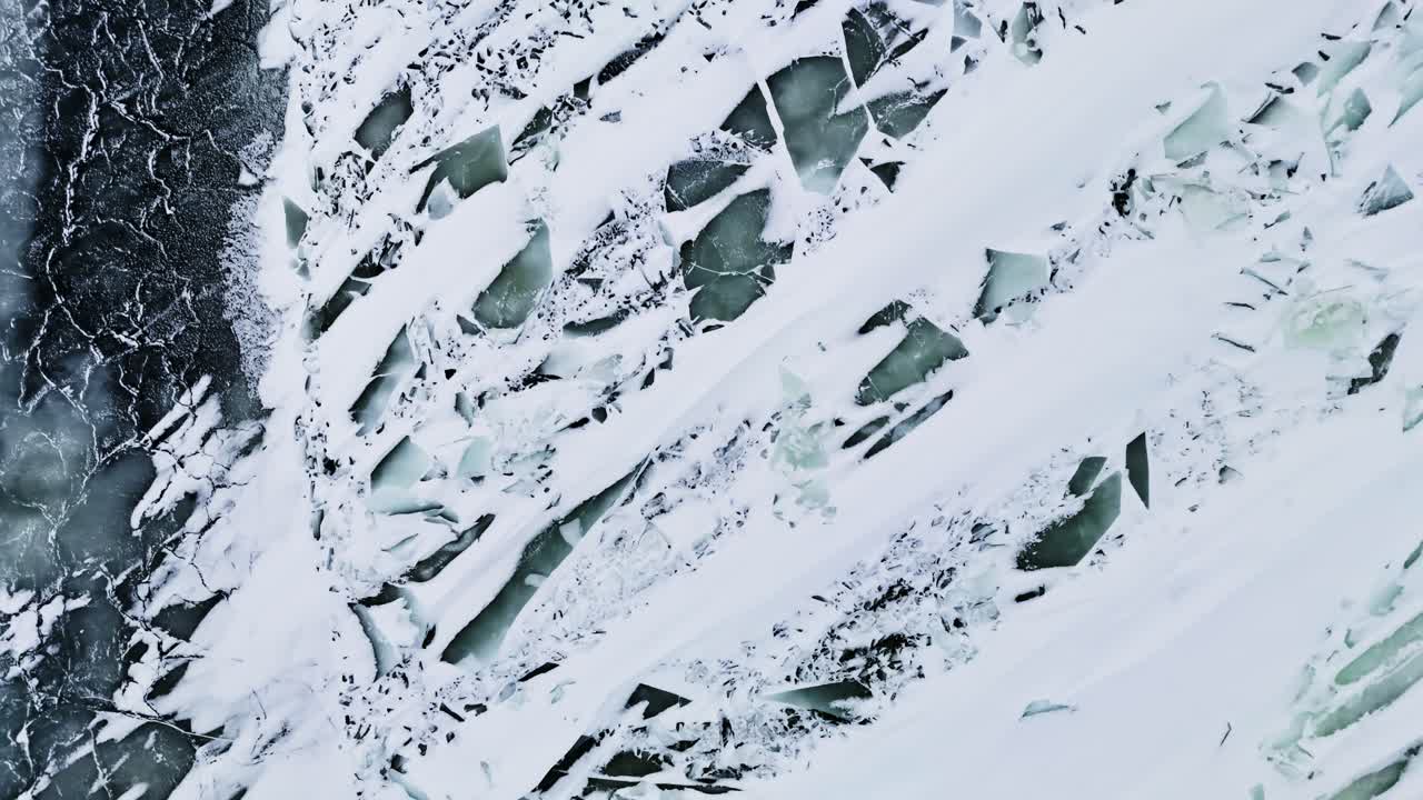 Primo piano da drone di ghiaccio marino congelato con striature di neve e crepe turchesi, che creano una texture invernale astratta e un motivo costiero drammatico