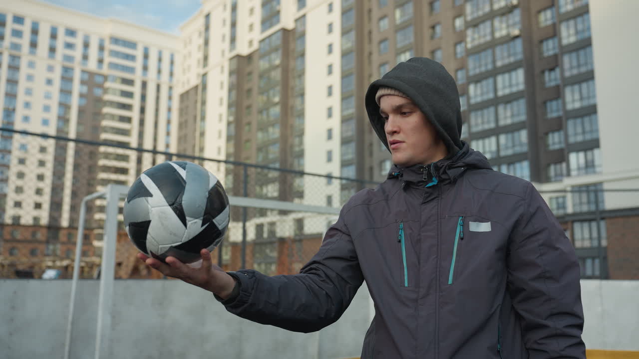 야외 스포츠 경기장에서 서 있는 후디를 입은 남자가 집중력과 집중력으로 손에 축구공을 회전시키고, 배경에는 도시 고층 건물이 보인다.