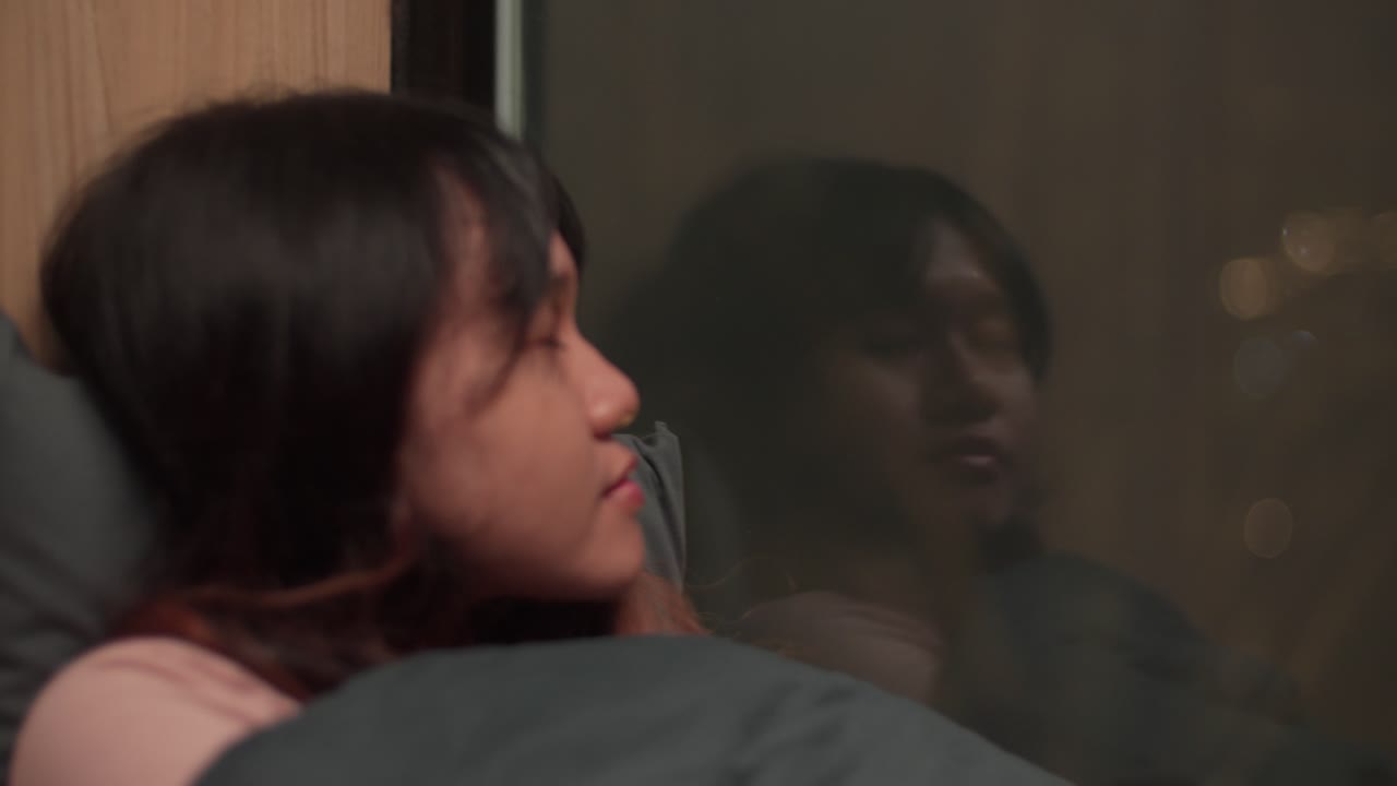 A girl with dark hair lying in bed at night, gazing out the window with a soft reflection