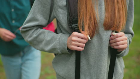 close-up of woman gripping backpack straps as she hikes through a lush forest, and jeans, with long hair flowing over her shoulders, accompanied by a friend in green shirt softly blurred behind