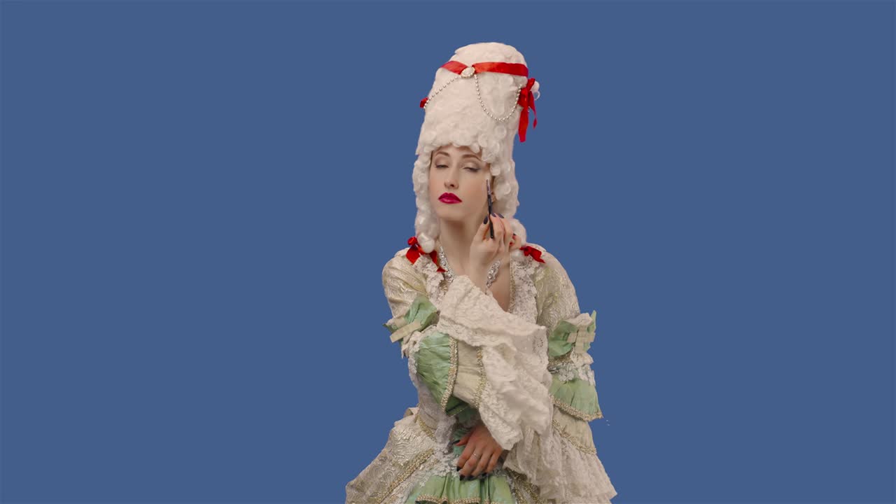 색 빈티지 렌즈 드레스와 위그를 입은 궁정 여인의 초상화, 브러쉬로 메이크업을 하고 있는 젊은 여성이 블루 스크린 배경으로 스튜디오에서 포즈를 취하고 있다.