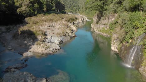 Beautiful-pristine-blue-clear-Pelorus-river,-New-Zealand-with-waterfall,-rocks-and-native-lush-forrest-in-background---Aerial-Drone