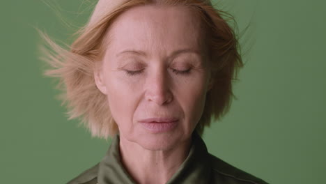 close up view of blonde mature woman wearing green shirt with closed eyes while the wind move her hair on green background, then she looks at camera