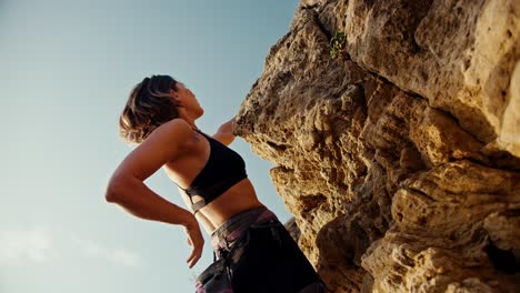 shooting from below: a blonde girl in a black top and black pants, using a special harness, climbs a rock and takes sports chalk out of her small bag for more confident climbing up the rock