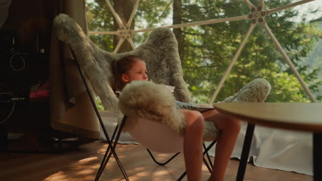 pensive little girl watches birds outside window swinging legs while sitting in fur chair. kid studies nature watching from indoors. accommodation in glamping