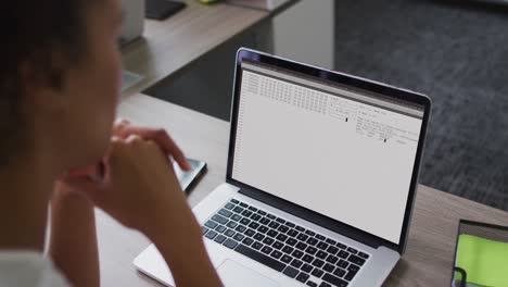 Mixed-race-woman-sitting-at-desk-watching-coding-data-processing-on-laptop-screen