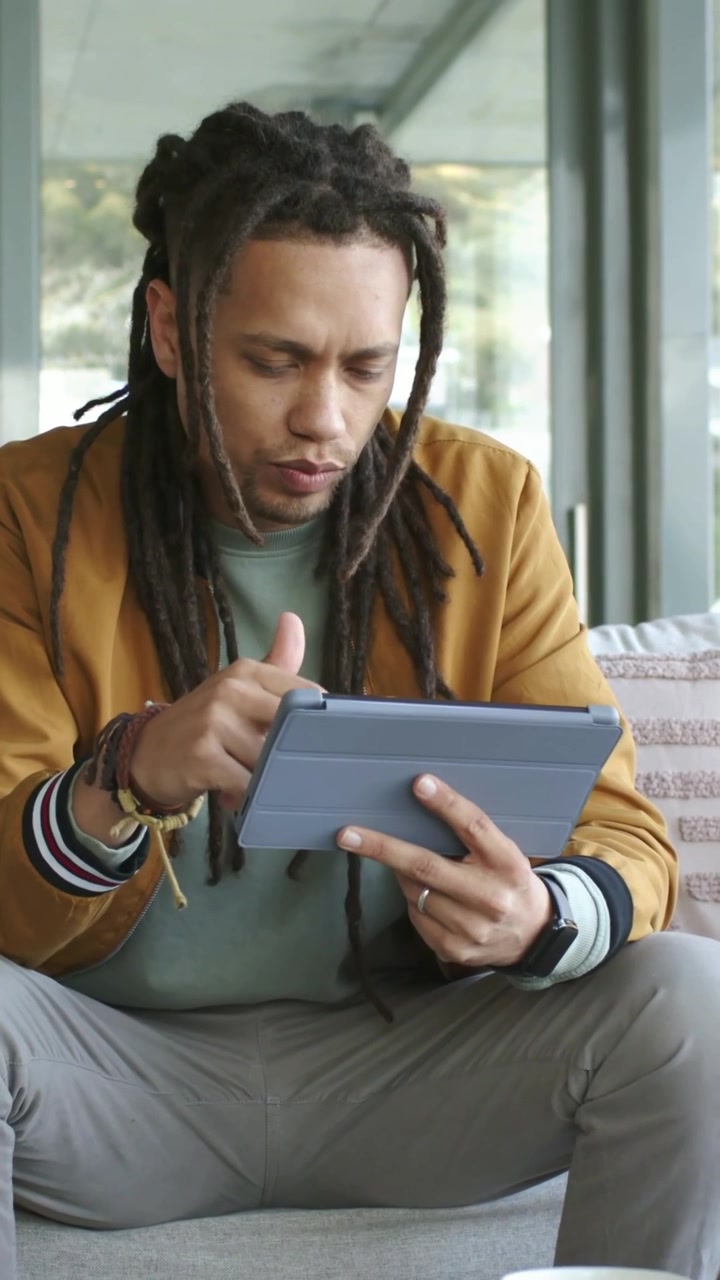 Vertical video: Tapping dreadlocked man in mustard jacket on sofa holding tablet, swiping to decide
