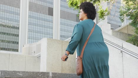 Happy-plus-size-biracial-woman-walking-on-stairs-with-smartphone-in-city