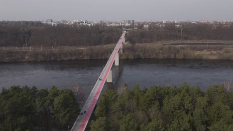 Drone-approaches-a-busy-pedestrian-bridge-over-a-river-with-a-city-in-the-distance