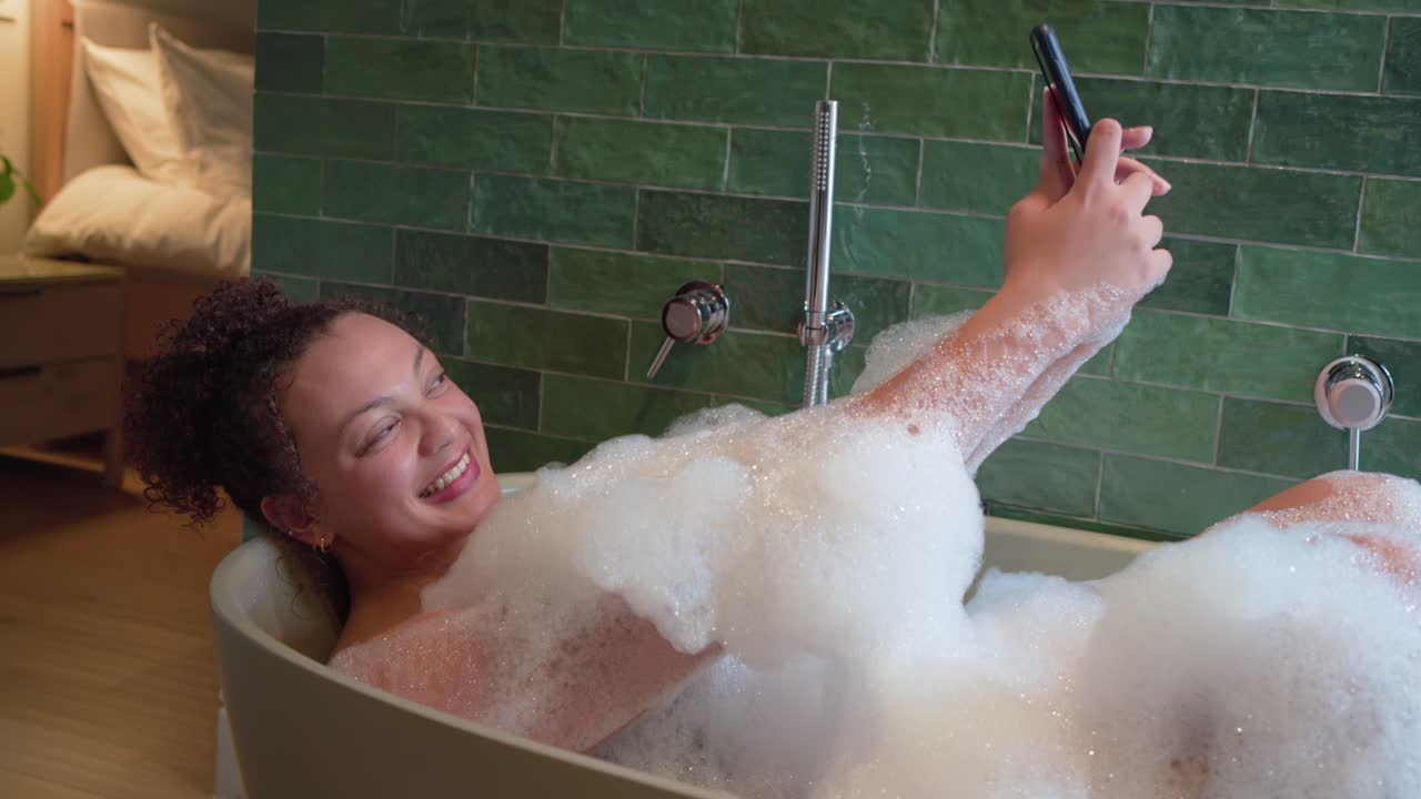 Afroamerikansk kvinde ligger i badekar og trykker på telefonen, mens hun tager selfies i skummet