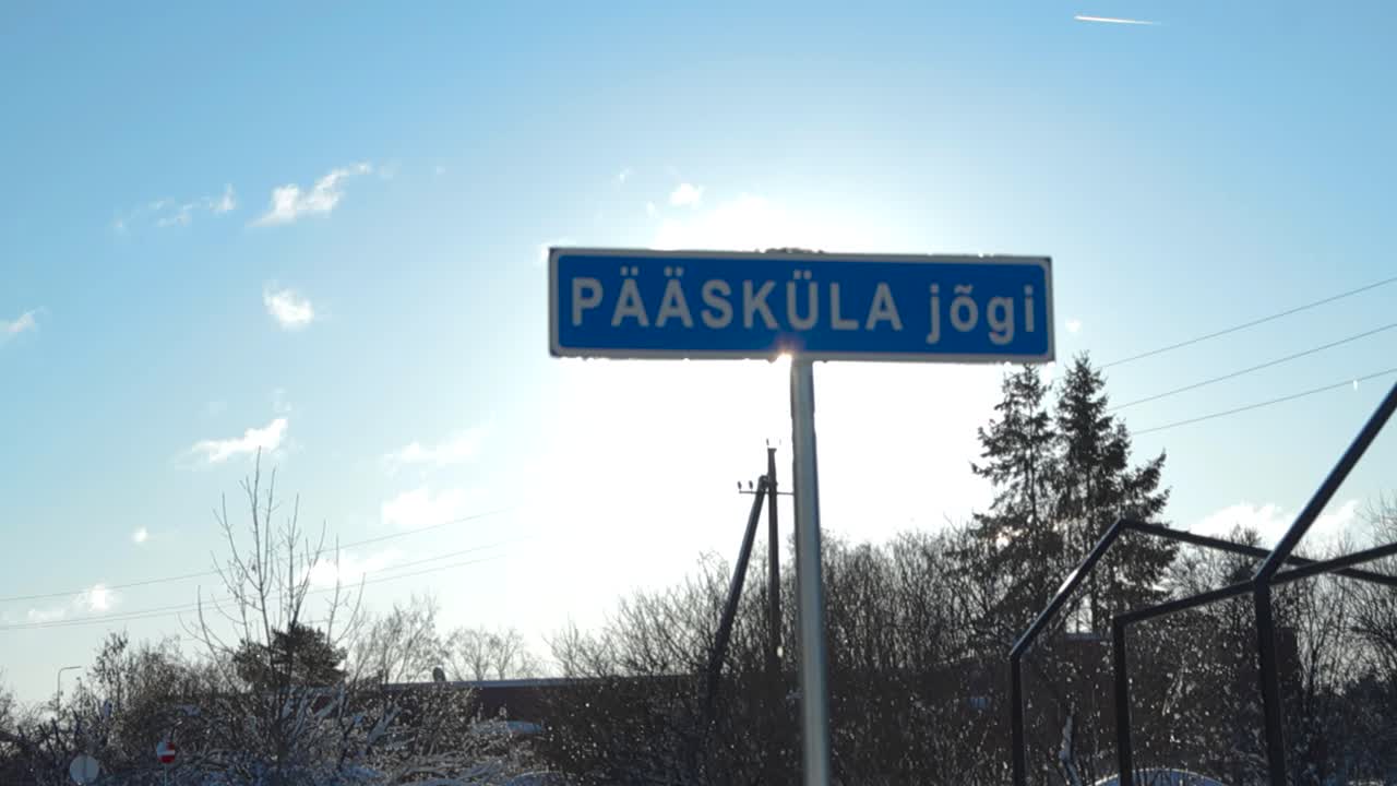 Gorgeous slowly panning footage of a blue colored sign that says Pöääsküla river in Estonian white letters on it. Sun is shining behind the sign and footage reveals fluffy white snow in the background