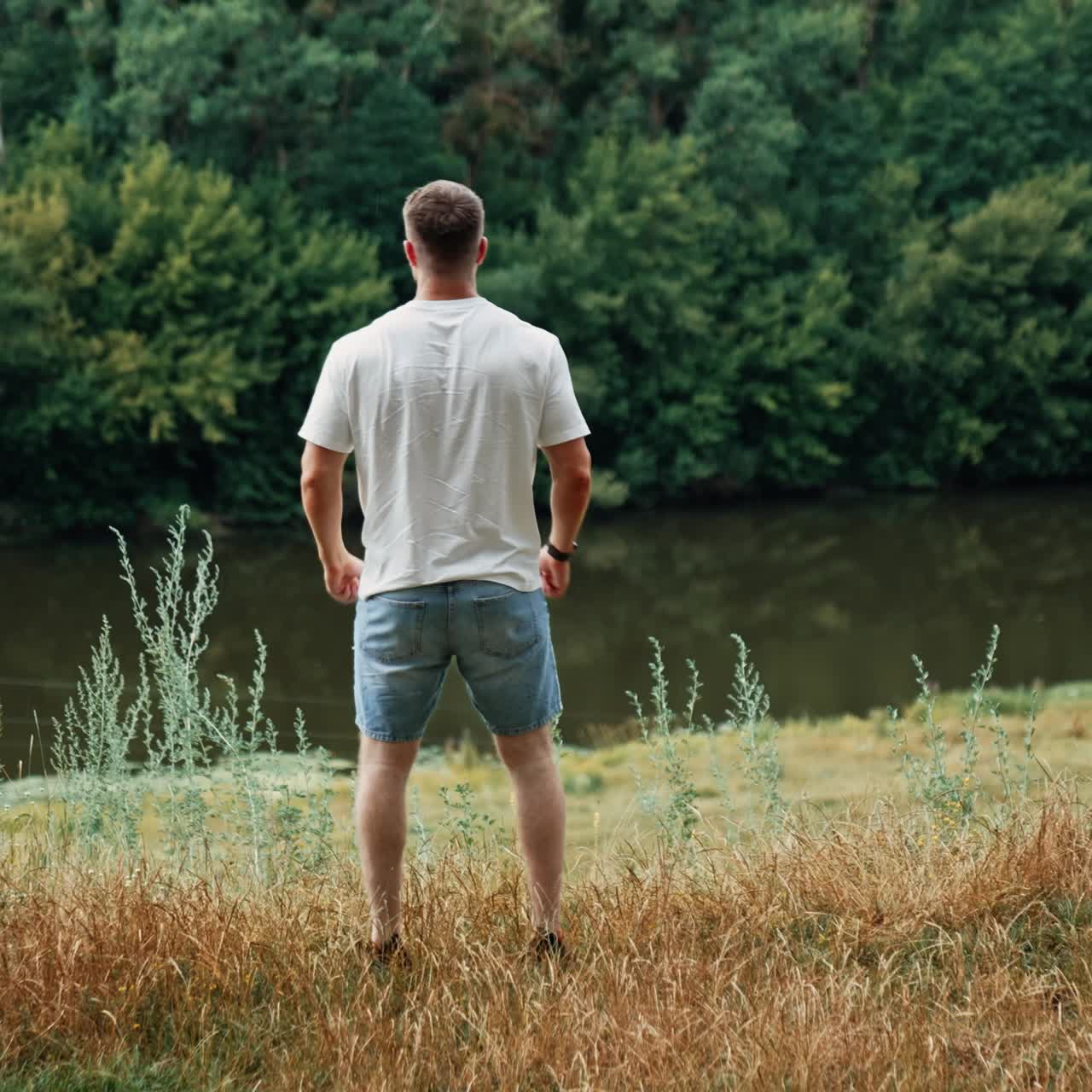Rear view of a strong muscular man in white t-shirt and jeans shorts. Man raises hands rejoicing life and free time in nature