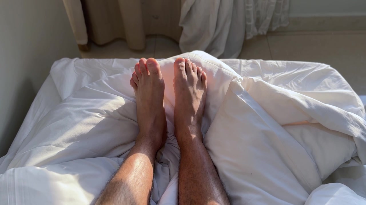 Bare male feet resting comfortably on white bed sheets and soft pillows. Bright morning sunlight shines through the bedroom curtains, creating a warm, relaxing atmosphere during a lazy weekend morning