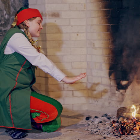 beautiful elf is getting warm while sitting near the fire at fireplace. female elf in fairy costume rubs her cold hands close to the fireside. christmas atmosphere indoors.