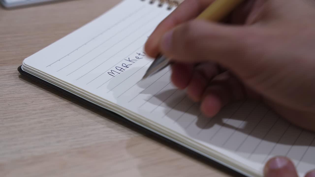 Nærbillede af en hånd, der skriver nummererede trin under titlen "markedsføringsplan" i en spiralnotesbog på et skrivebord, illustrerende forretningsplanlægning, strategiudvikling og organiserede arbejdsgange